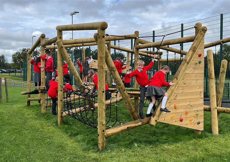 A group of children wearing red jumpers are climbing over a wooden structure with a variety of different challenges. From balance beams to rope tunnels, the wooden structure is quite big.